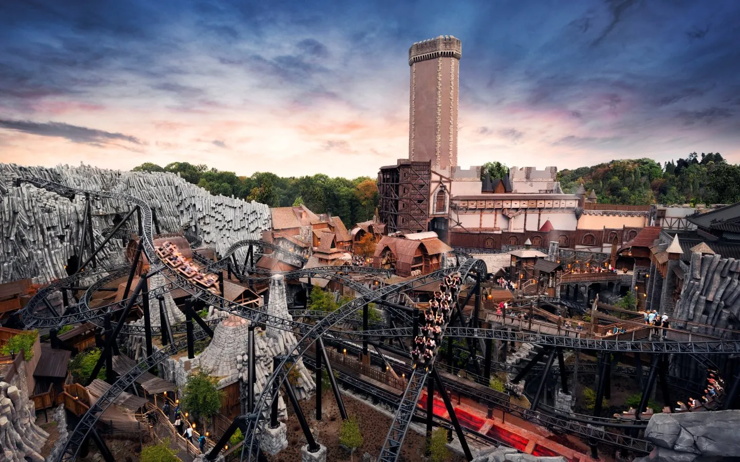Panoramic view of Phantasialand theme park featuring dramatic roller coaster tracks weaving through a medieval-themed village, with a prominent tall tower structure in the background against a dramatic sunset sky, showcasing both thrilling rides and detailed themed architecture throughout the park landscape